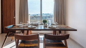 Una mesa de comedor dispuesta con platos, vasos y cubiertos, ubicada frente a un ventanal con cortinas que da a un balcón.
