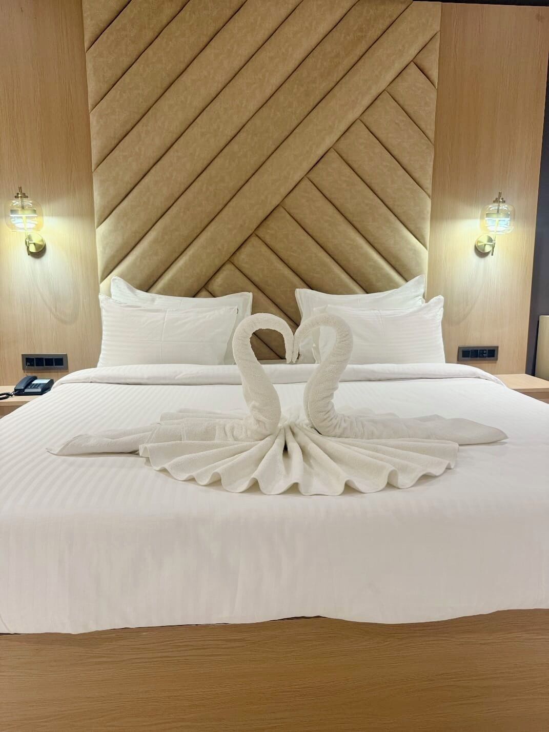A hotel room with a large bed, a headboard with a geometric pattern, two wall-mounted light fixtures, and a swan-shaped towel arrangement on the bed.