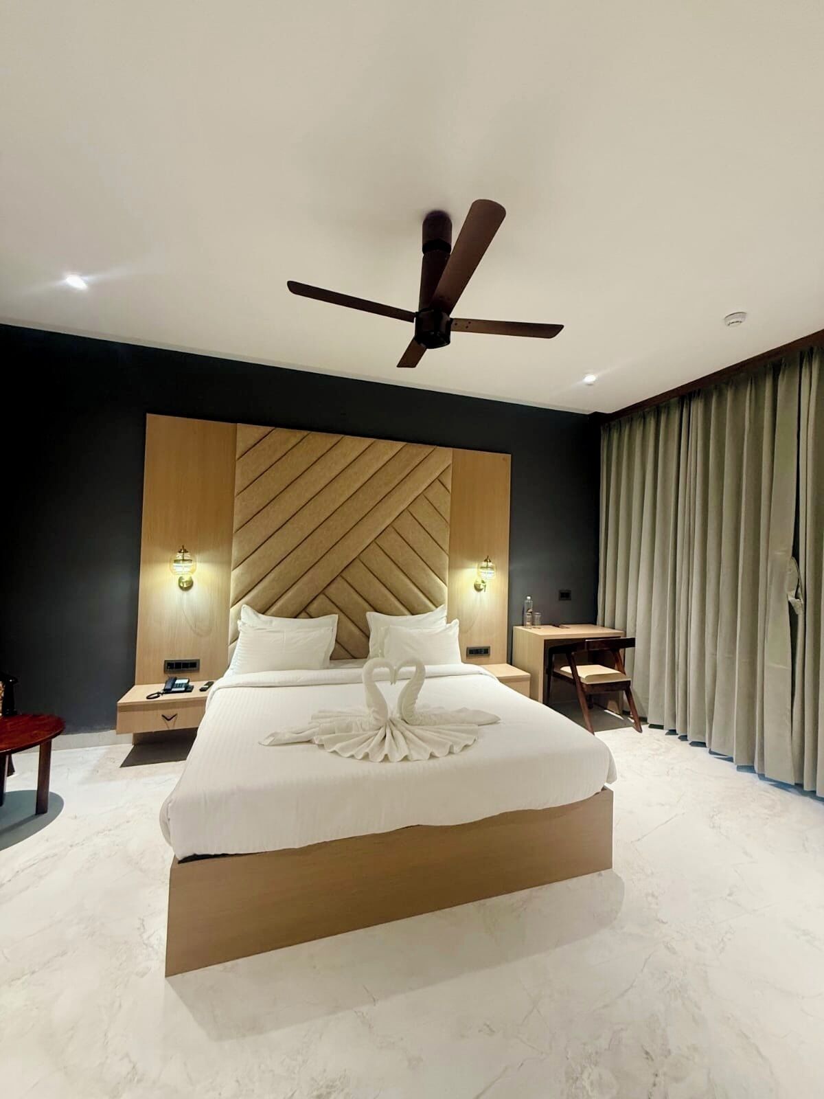 Quarto de hotel moderno com uma cama grande, ventilador de teto, cabeceira de madeira, abajures, escrivaninha e uma cadeira.