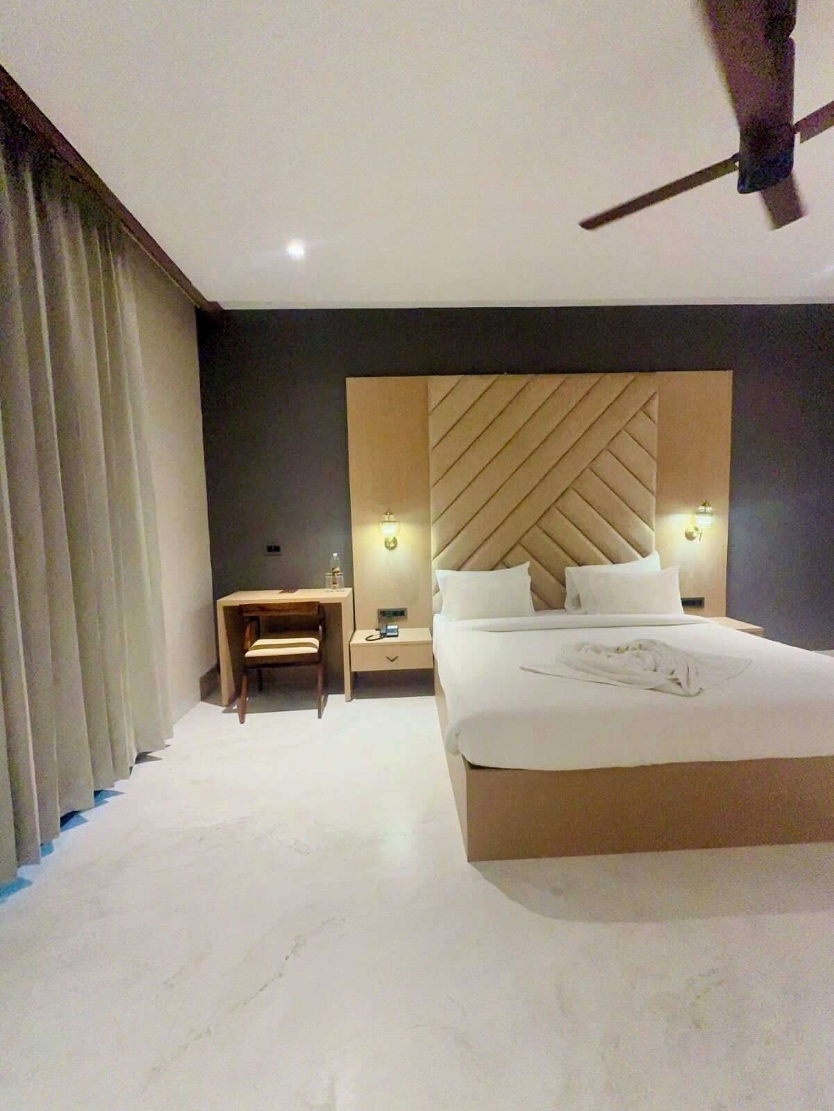 A hotel room with a large bed, a desk, a chair, and a ceiling fan.