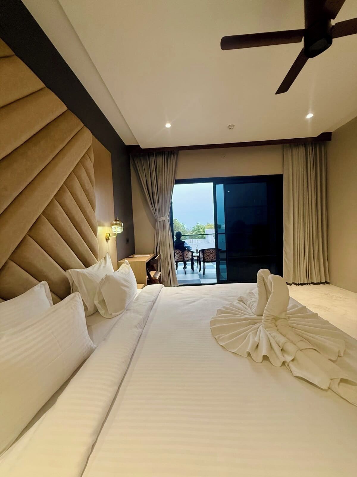Une chambre d’hôtel avec un grand lit, un ventilateur de plafond, une vue sur l’extérieur et un coin repas visible à travers la porte vitrée.