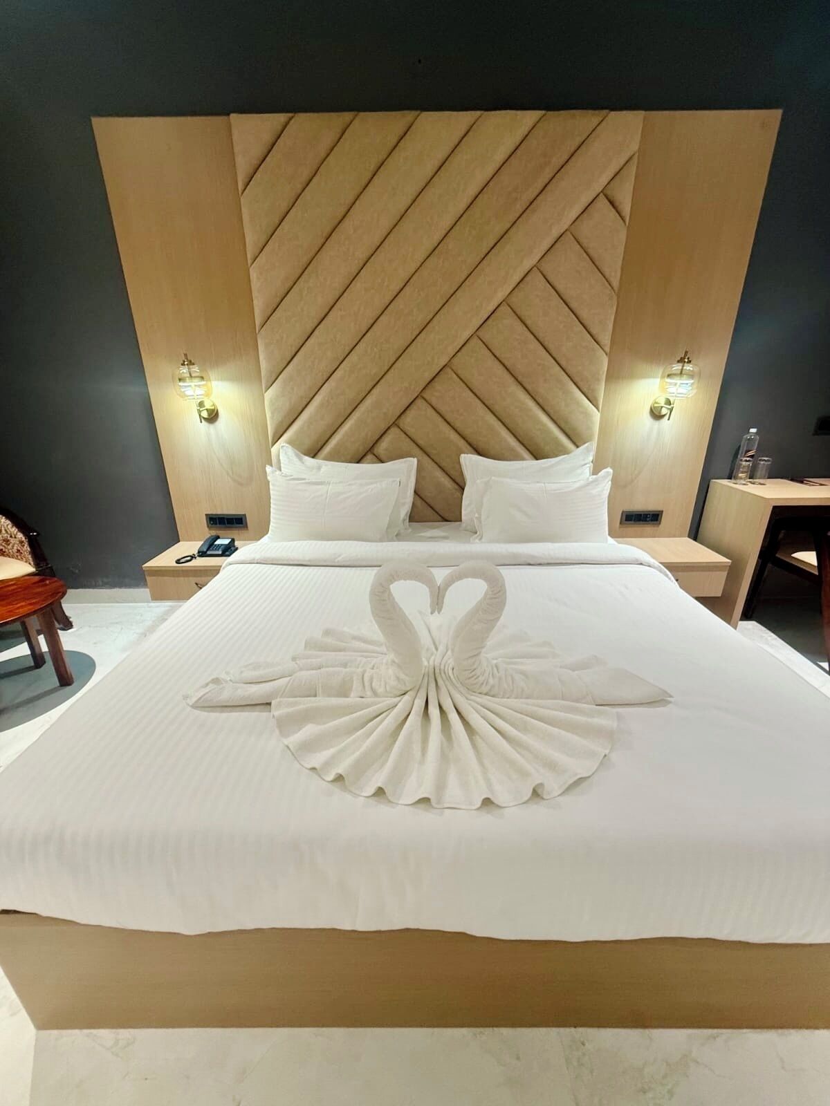 Hotellihuone, jossa on suuri sänky, puinen pääty, kaksi yöpöytää valaisimineen ja koristeellinen joutsenenmuotoinen pyyhe sängyllä.
