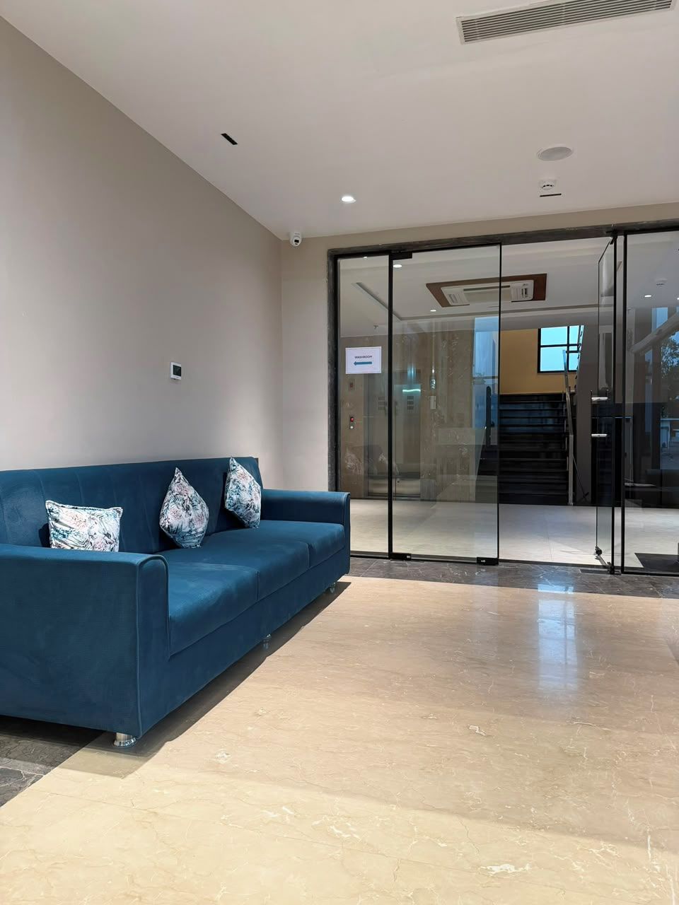A modern lobby with a blue sofa, glass doors leading to a staircase, and a sign indicating a waiting area.