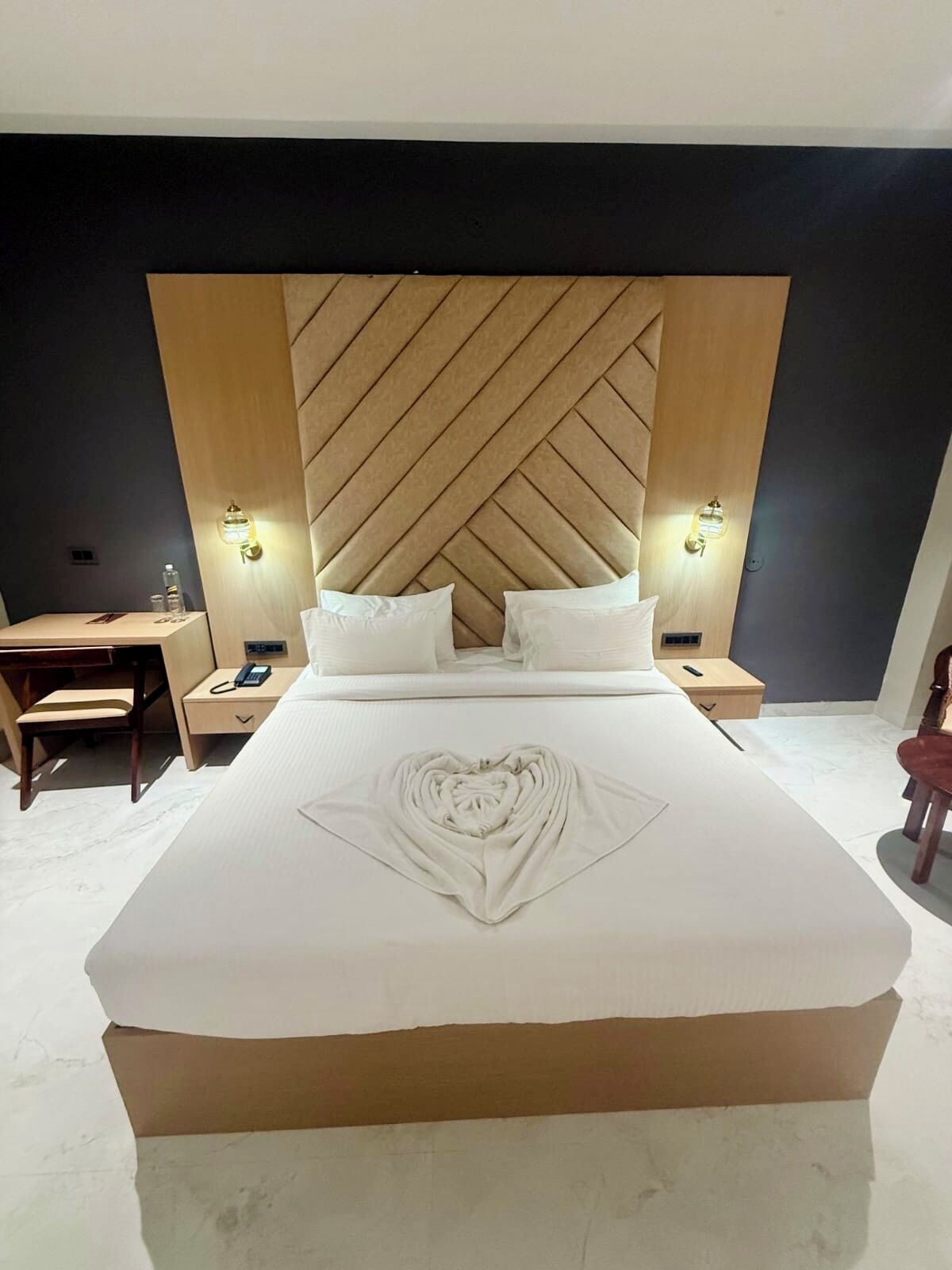 A hotel room with a large bed, a wooden headboard, bedside lamps, a desk, and a chair.