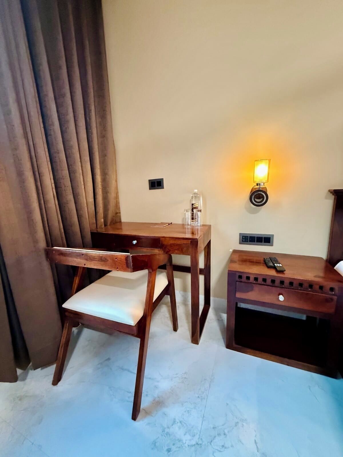 A room with a wooden desk, a chair, a bedside table with a remote control, and a wall-mounted light fixture.