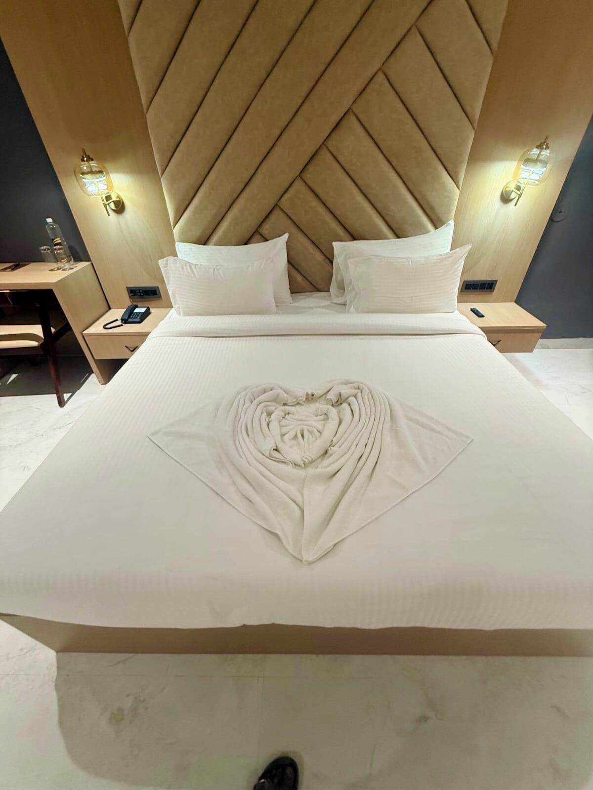 A hotel room with a large bed, a headboard with a diamond pattern, two bedside tables with lamps, and a neatly made bed with a heart-shaped towel design.