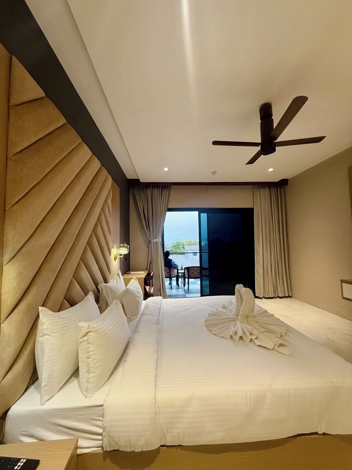Een hotelkamer met een groot bed, een plafondventilator, een bureau met stoel en uitzicht op de omgeving.