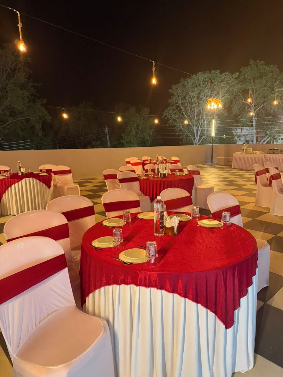 A banquet hall with round tables, white chairs with red sashes, and string lights.