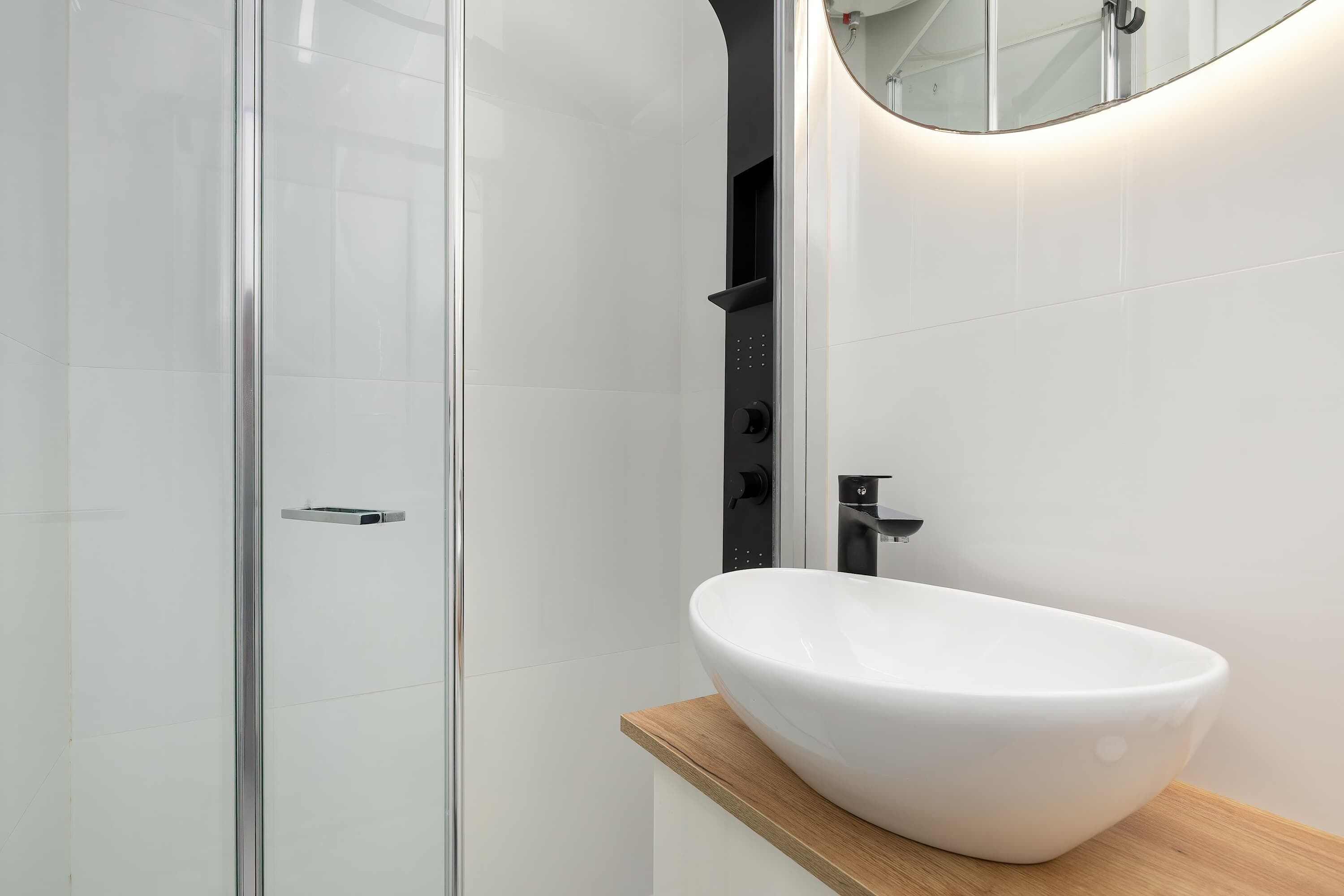 A modern bathroom with a glass shower enclosure, a white sink on a wooden countertop, and a mirror with a round frame.