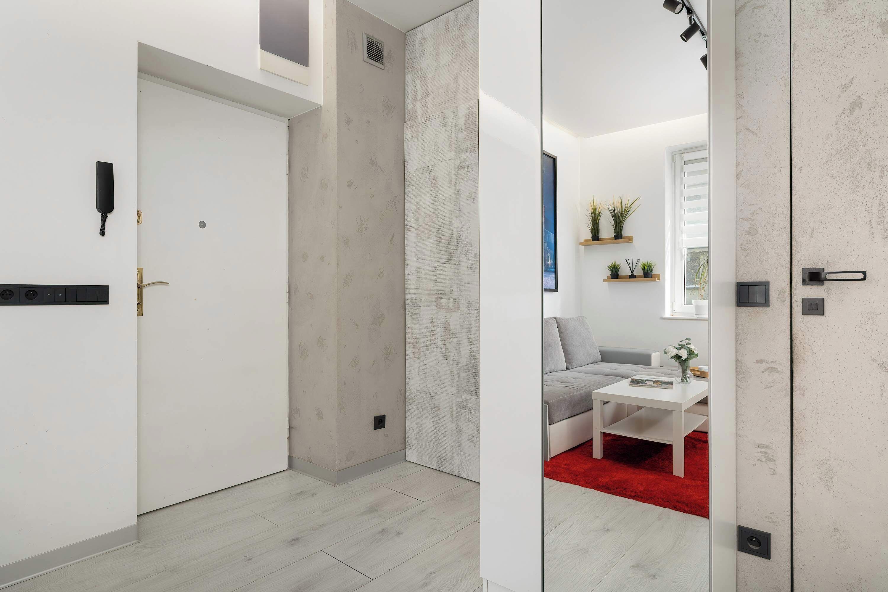 A modern hotel room with a white door, a red carpet, a grey sofa, and a white coffee table.