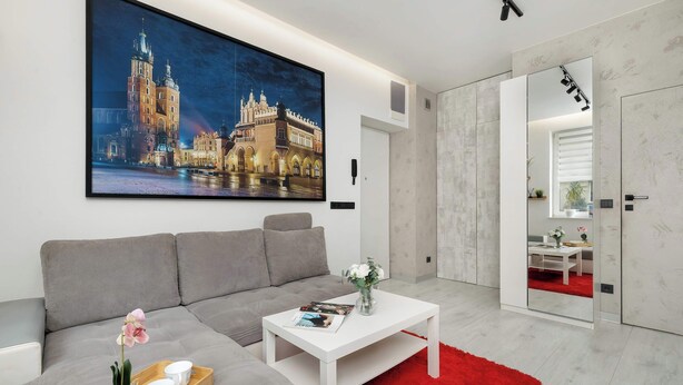 A modern living room with a grey sofa, a white coffee table, and a red rug. A large framed picture of a building is on the wall. - Cozy Studio for Two | 800 m from Blonia Krakowskie (Kraków)