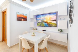 Un espace repas moderne avec une table et des chaises blanches, un grand téléviseur à écran plat fixé au mur au-dessus, et une porte sur le côté gauche.