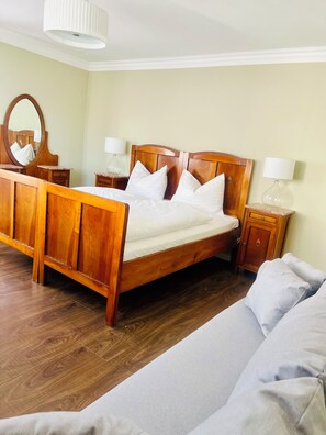 A wooden bed with white bedding, a nightstand with a lamp, a mirror, and a wooden floor.