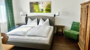 Een net opgemaakt bed met witte lakens, een met groen bekleden stoel, een houten kast en een abstract schilderij aan de muur.