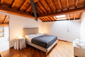 A bedroom with a wooden ceiling, a bed with a grey quilt, bedside lamps, a wardrobe, and a small round table with a lamp. - Cozy penthouse in the heart of via Mazzini (Verona)