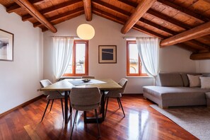 A room with a wooden ceiling, a dining table with chairs, a sofa, and two windows with curtains. - Cozy penthouse in the heart of via Mazzini (Verona)