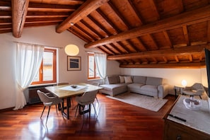 Ein geräumiges Zimmer mit Holzbalkendecke, einem Esstisch mit Stühlen, einem Sofa und einem kleinen Tisch mit Lampe.