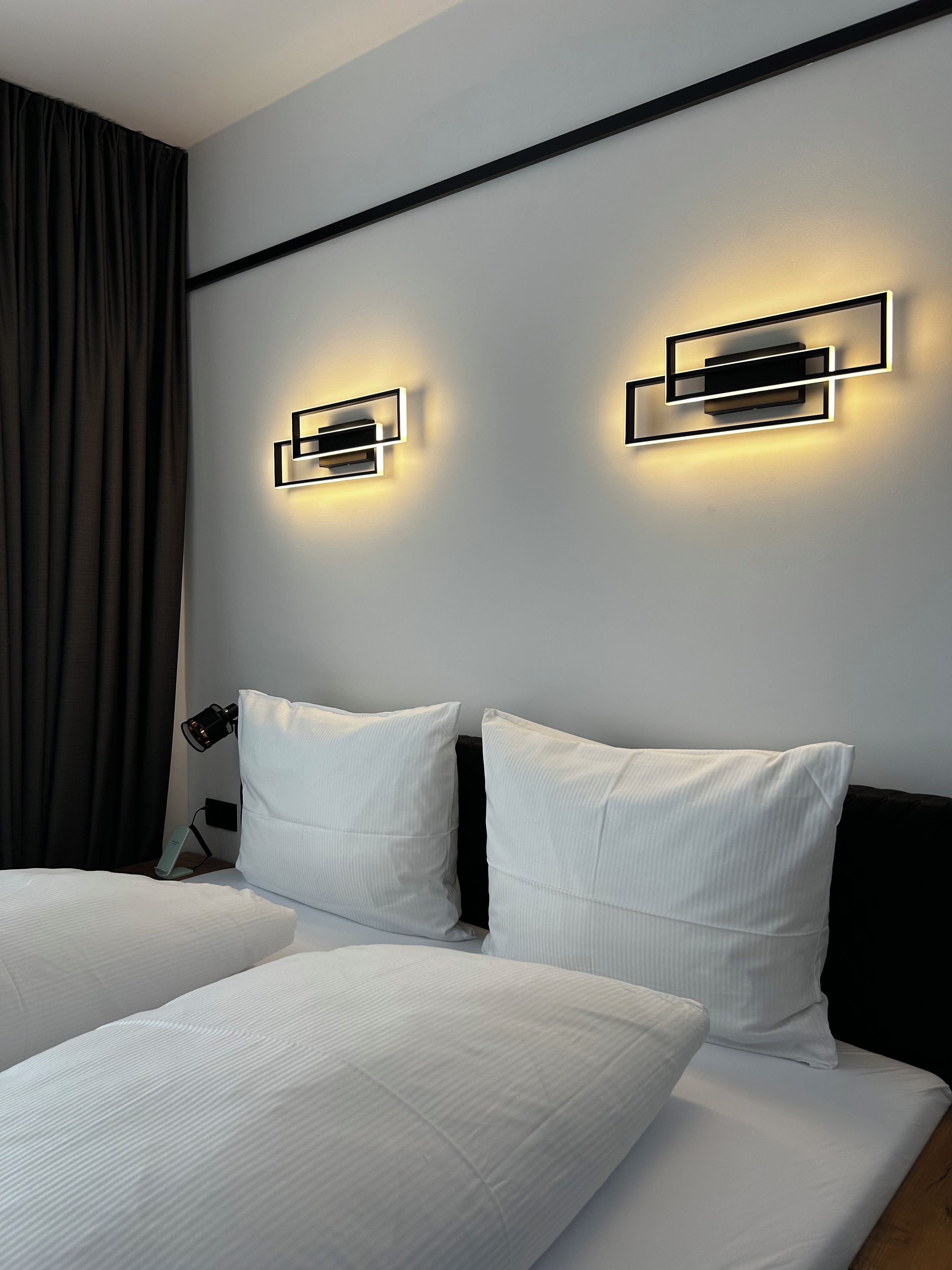 A hotel room with two beds, a nightstand, a lamp, and wall-mounted lighting fixtures.