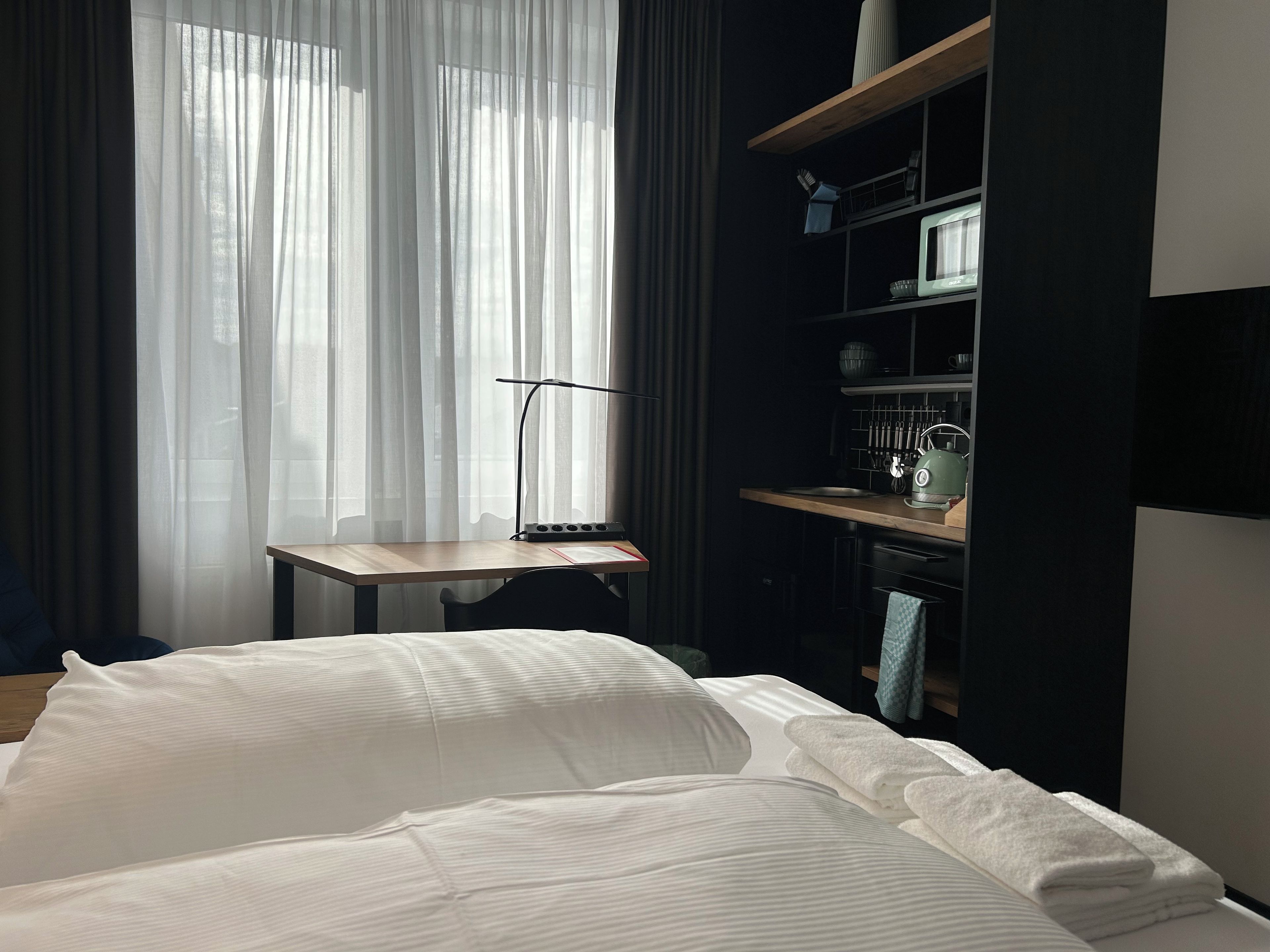 A hotel room with a bed, a desk, a shelf with kitchenware, and a window with sheer curtains.