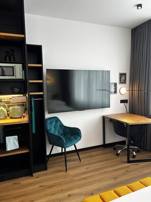 A modern room with a flat-screen TV, a teal chair, a wooden desk, and a kitchen area with a microwave and kettle. - The Loft (Münster)