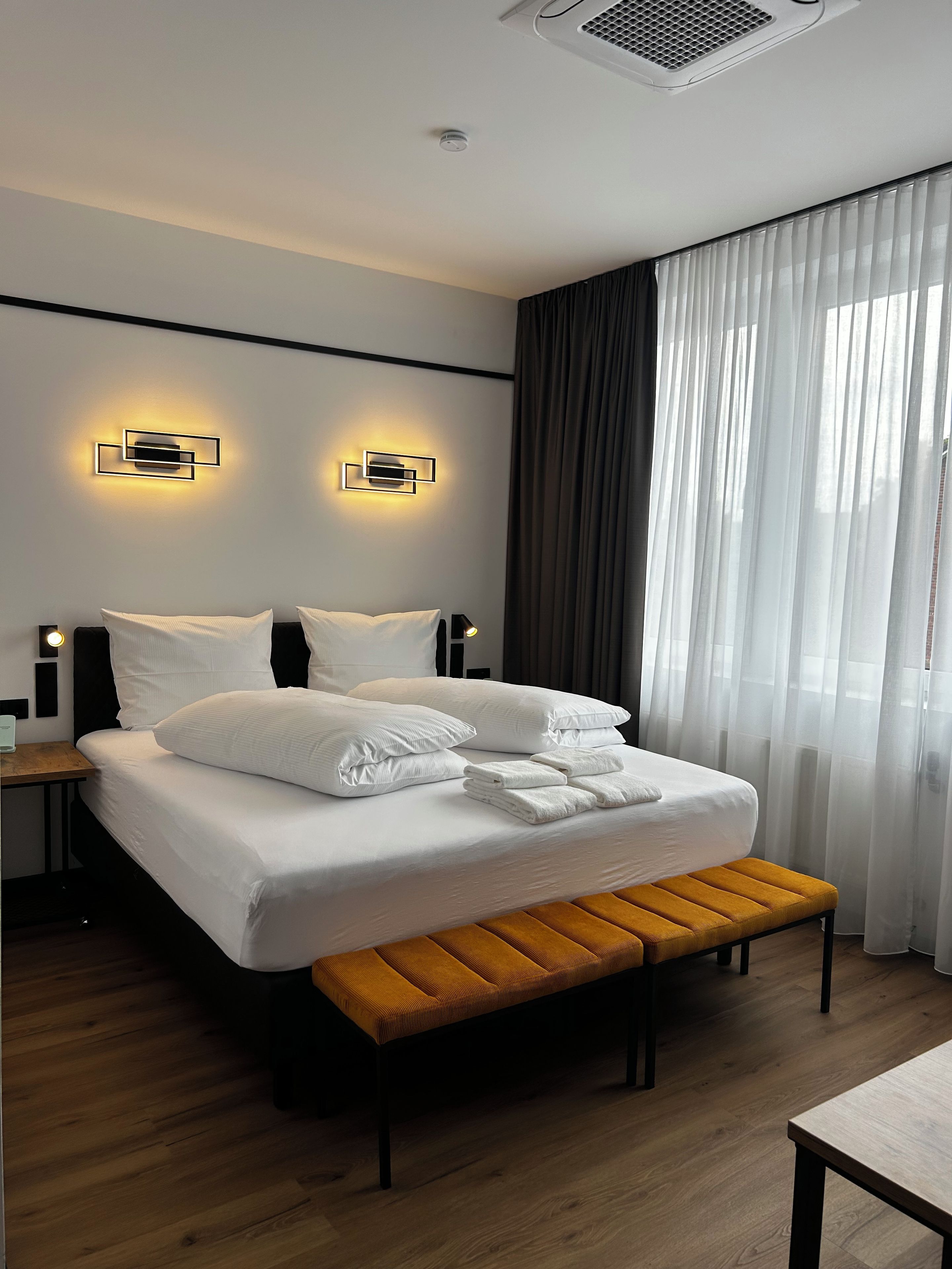 A modern hotel room with a large bed, bedside tables, a bench, and a window with sheer curtains.