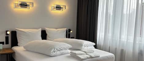 A modern hotel room with a large bed, bedside tables, a bench, and a window with sheer curtains.