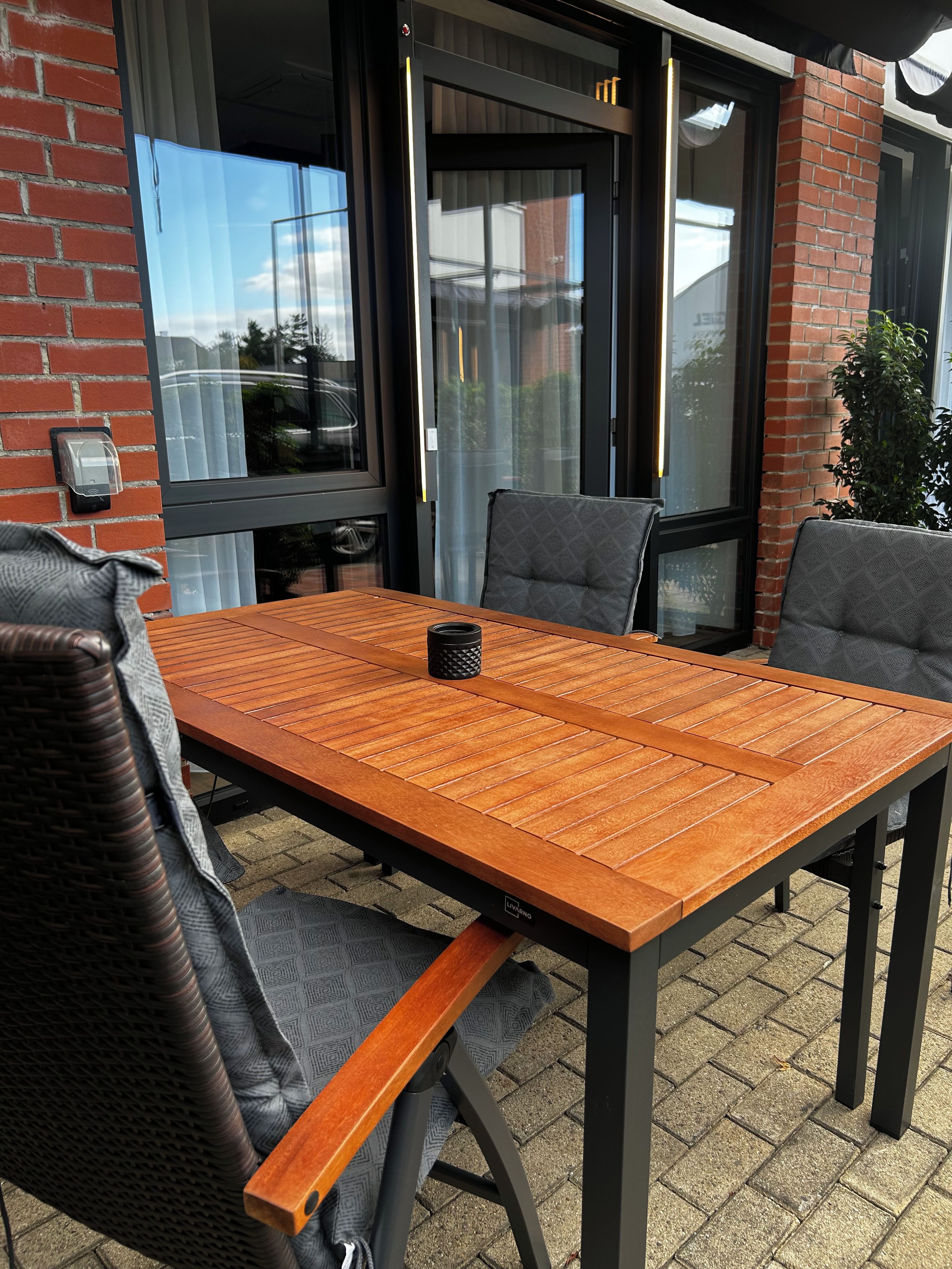 Un patio exterior con una mesa y sillas de madera, un muro de ladrillo y una puerta de cristal.