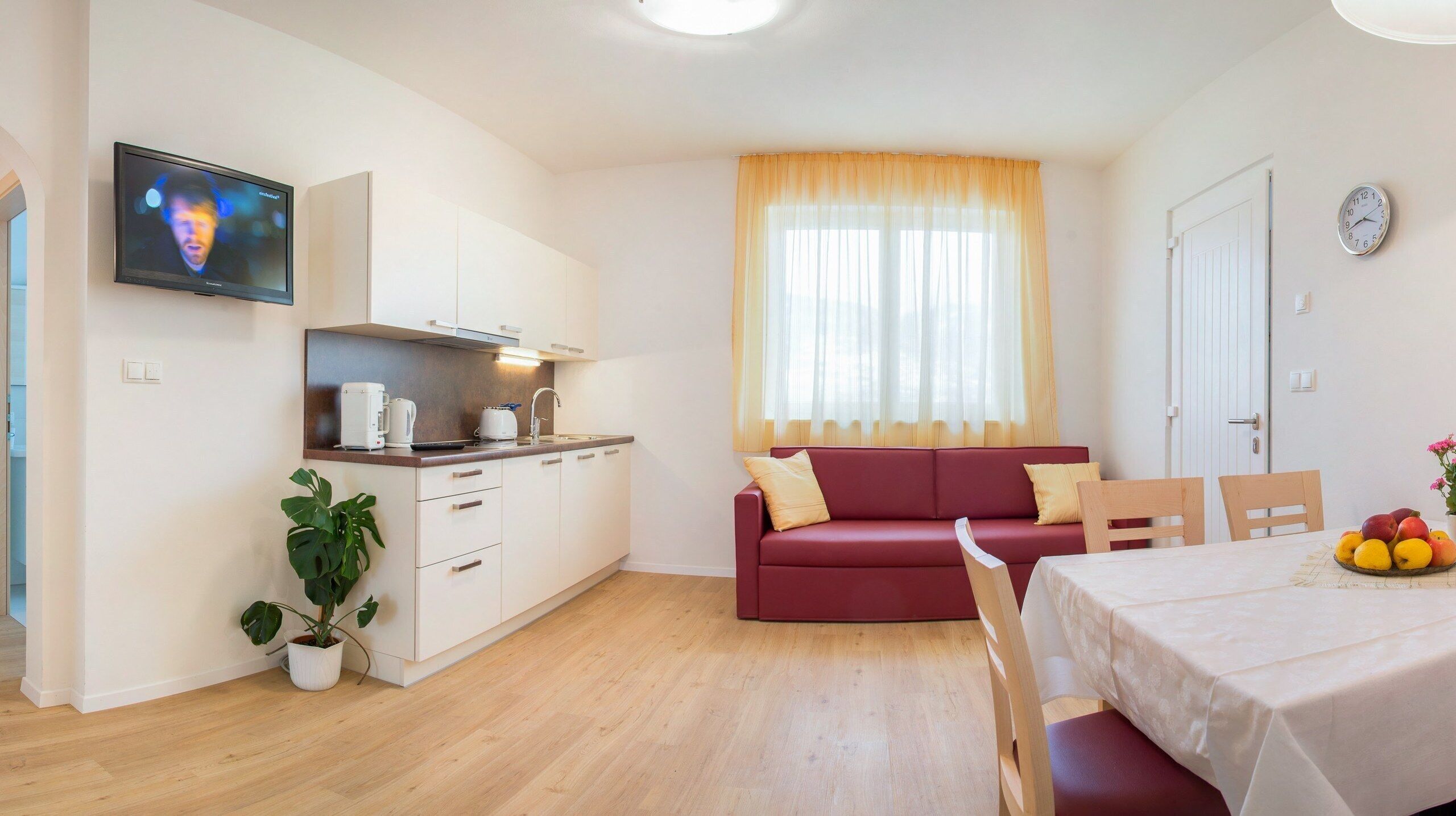 Un salon moderne avec un coin cuisine, un canapé rouge, une table à manger ornée de fruits et un téléviseur fixé au mur.