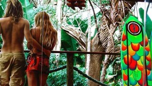Un couple se tient sur une terrasse en bois, avec une vue sur une végétation luxuriante et une planche de surf colorée appuyée contre une structure.