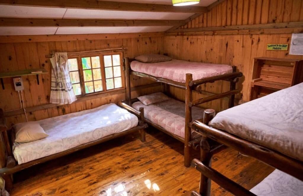 Una baita con letti a castello, pareti in legno, una finestra con tende e un cartello appeso al muro.