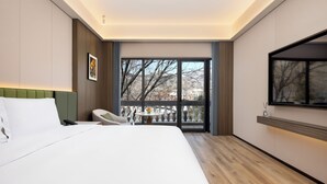 Una moderna camera d'albergo con un letto grande, un tavolino con una sedia, una TV a parete e un balcone con vista sugli alberi.