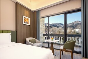 A hotel room with a bed, two chairs, a small table, a painting, and a balcony with a mountain view. - Fragrant Hill Empark Hotel (Beijing)