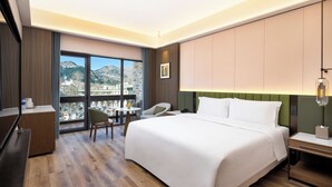 Una habitación de hotel con una cama grande, un escritorio, una silla, una mesita y vistas a montañas a través de amplios ventanales.