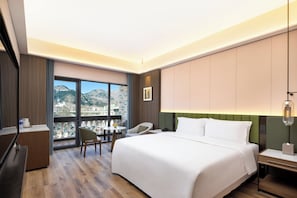 A hotel room with a large bed, a desk, a chair, a small table, and a view of mountains through large windows.