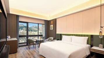 A hotel room with a large bed, a desk, a chair, a small table with a fruit bowl, and a view of mountains through the window.
