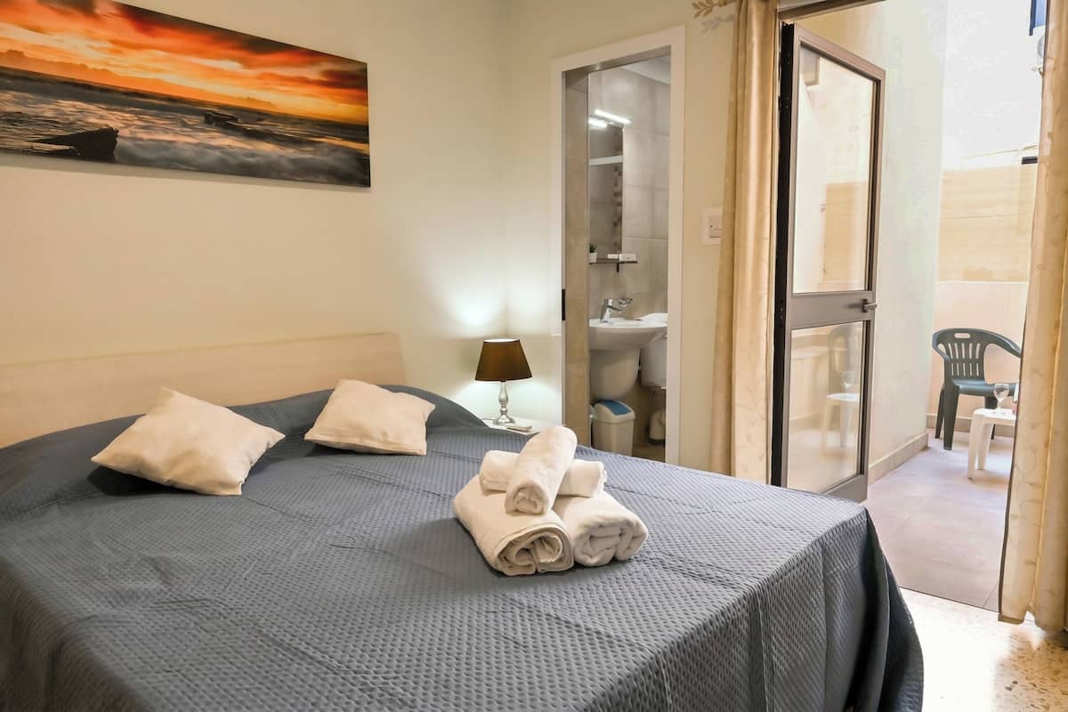 A hotel room with a bed, a painting, a bathroom visible through a glass door, and a balcony with chairs.