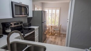 Een keuken met een magnetron, fornuis, koelkast en wastafel. Er is een eethoek met een tafel en stoelen.
