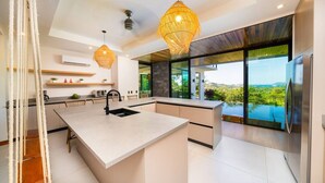 Een moderne keuken met een centraal eiland, hanglampen en uitzicht op een weelderig landschap door grote ramen.
