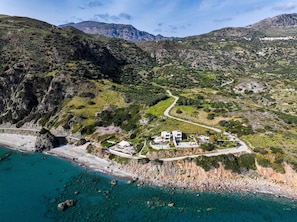 Luftaufnahme einer Küstenlandschaft mit kurviger Straße, felsigem Gelände und Gebäuden mit Schwimmbad.