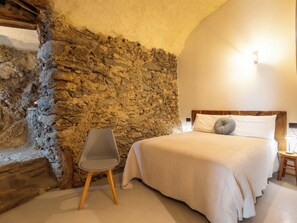 Ein Schlafzimmer mit Steinwand, einem Bett mit weißer Bettwäsche, einem grauen Sessel und einem Holzkopfteil.