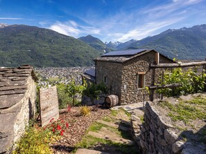 Ein Steinhaus mit kleinem Fenster, einer Solaranlage und einem Weinberg im Vordergrund.