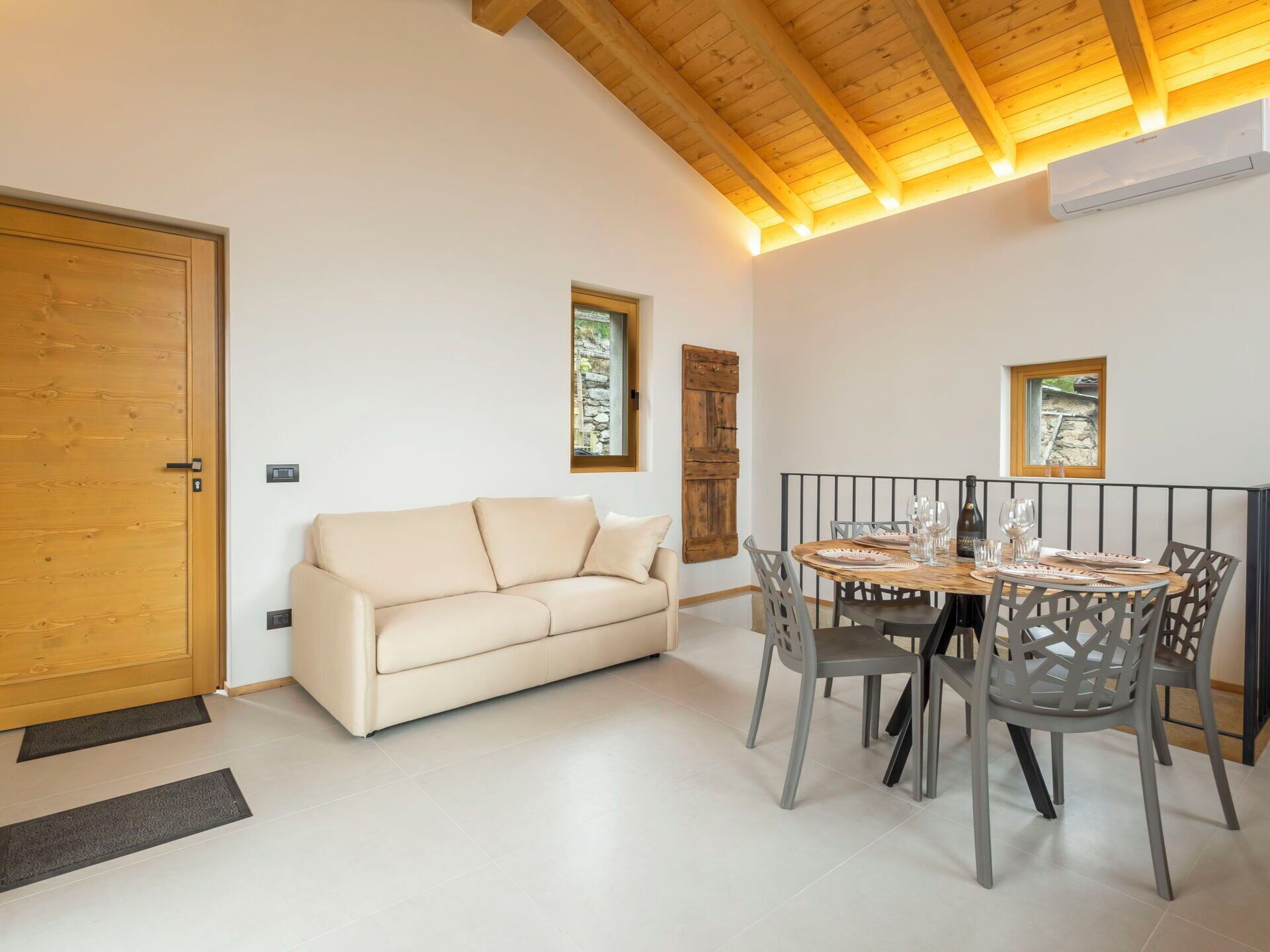 A room with a wooden door, a beige sofa, a dining table with chairs, and a window with a view.