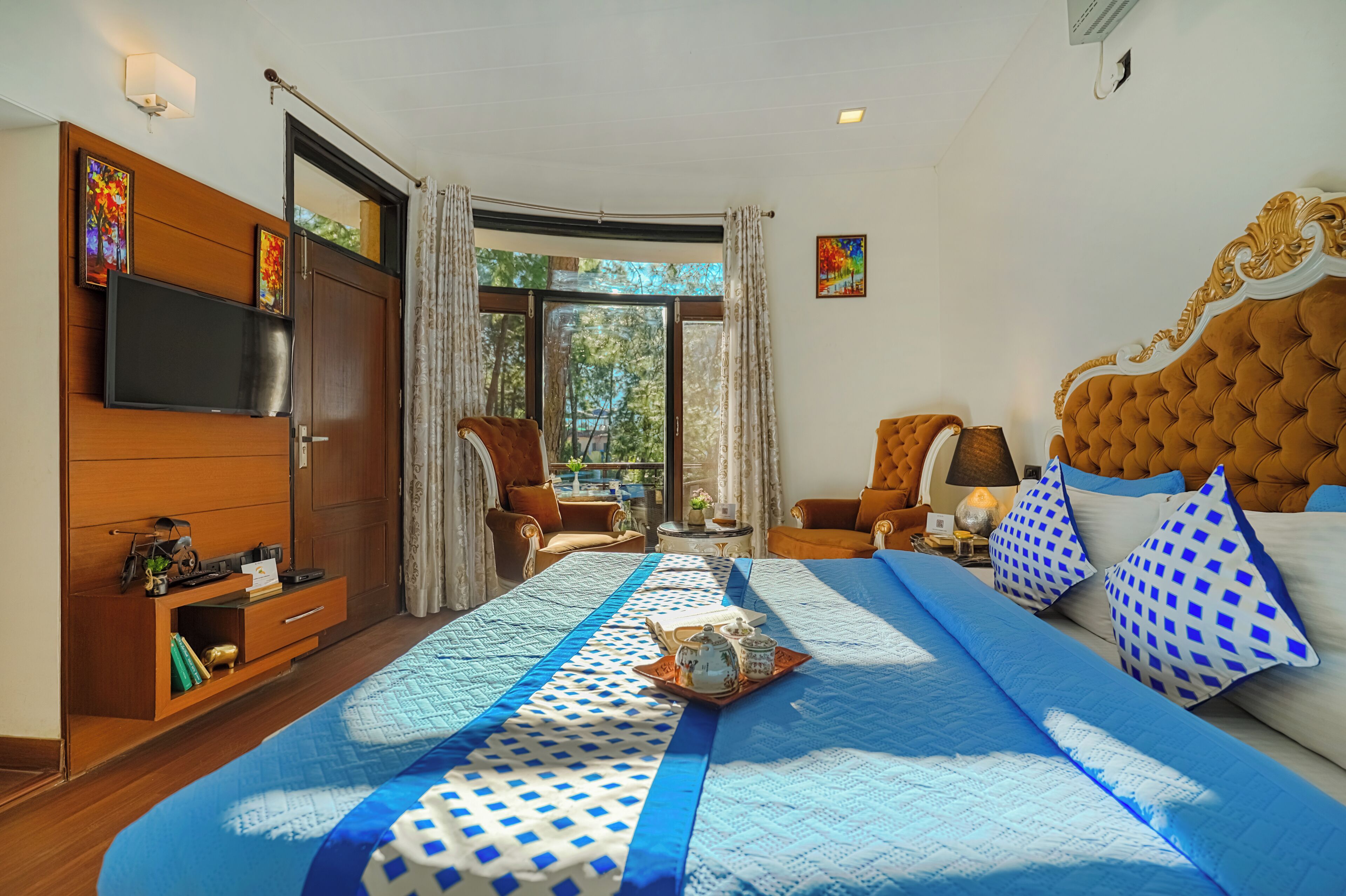 A hotel room with a large bed, a TV, a wardrobe, a seating area with a table, and a window with curtains.