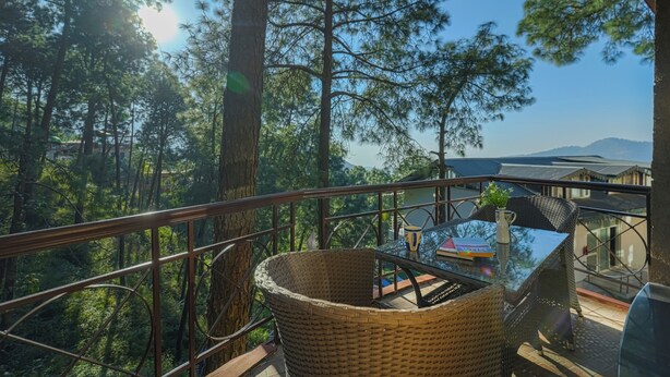 A balcony with a wicker chair, a glass table, and a view of a forest and mountains. - Alaya Stays 3BHK Golden Meadow Villa (Kasauli)
