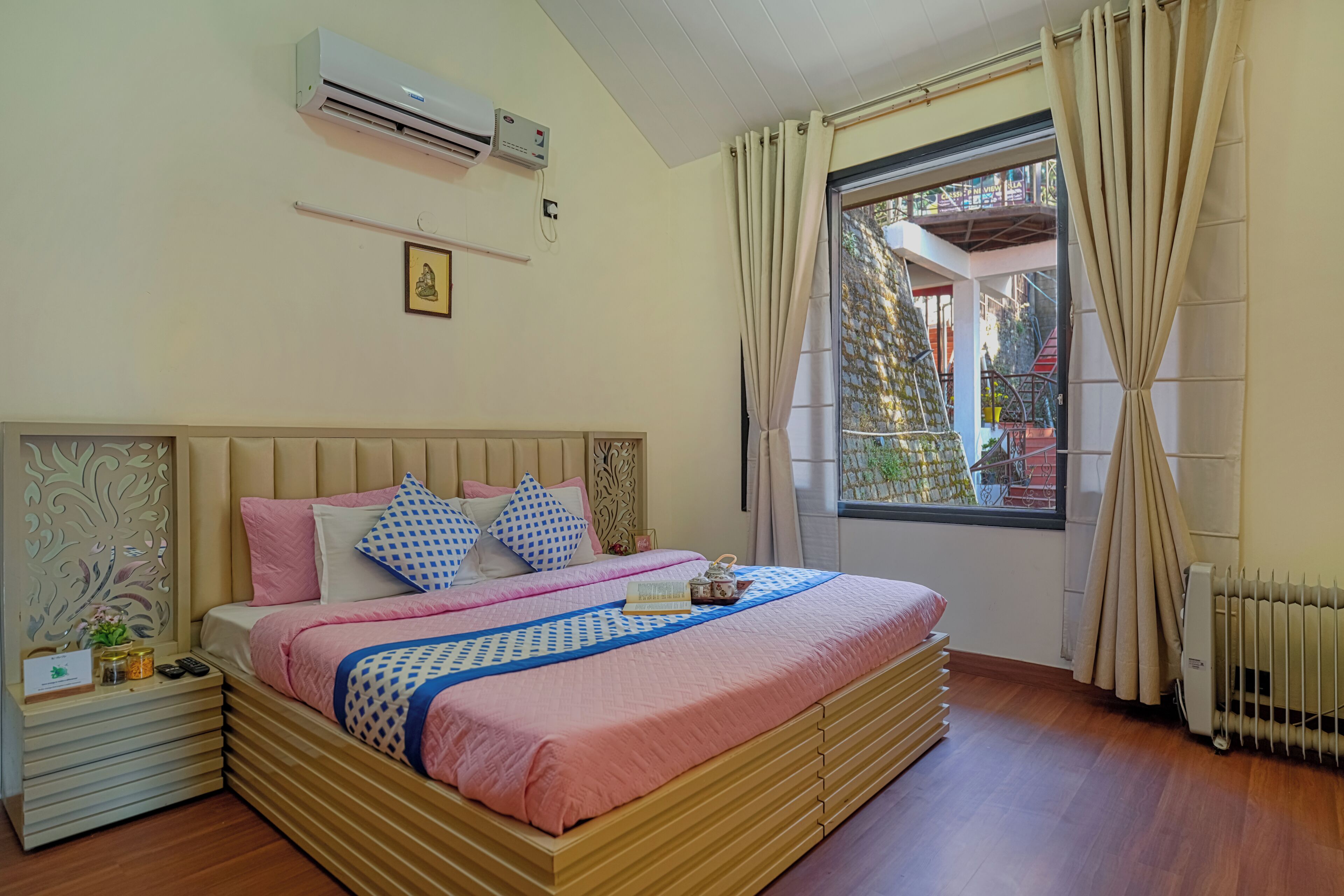 A bedroom with a bed, bedside table, air conditioning unit, window with curtains, and a radiator.