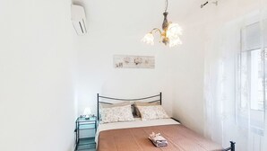 Uma cama de solteiro com estrutura de metal, uma mesa de cabeceira com abajur, um lustre e uma janela com cortinas finas.