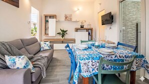 Sala moderna com sofá, mesa de jantar e cadeiras. Há uma área de cozinha com televisão e uma janela.