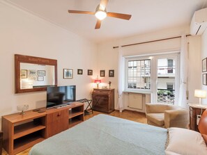 Une chambre à coucher comprenant un lit, une télévision, un bureau, une chaise, une fenêtre et un ventilateur de plafond.