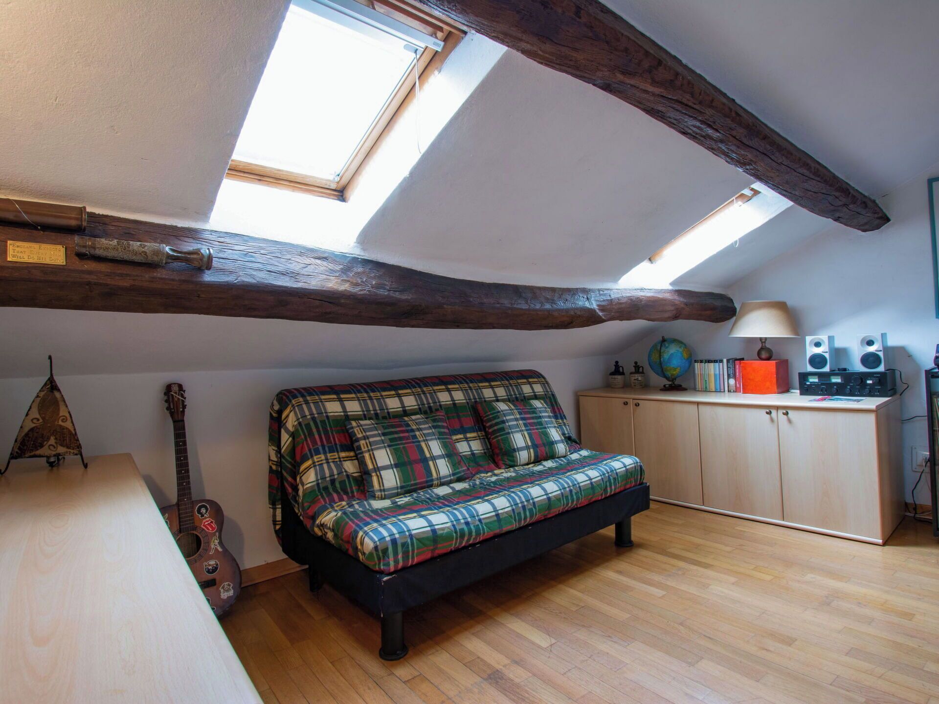 Ein Schlafzimmer im Loft-Stil mit schrägen Wänden, freiliegenden Holzbalken, einem Karosobsofa, einer Gitarre, einem Schrank und einem Schreibtisch mit Lampe und Lautsprechern.