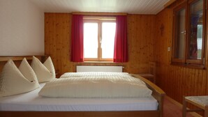 Quarto com painéis de madeira, uma cama, almofadas, uma janela com cortinas vermelhas e uma cadeira de madeira.